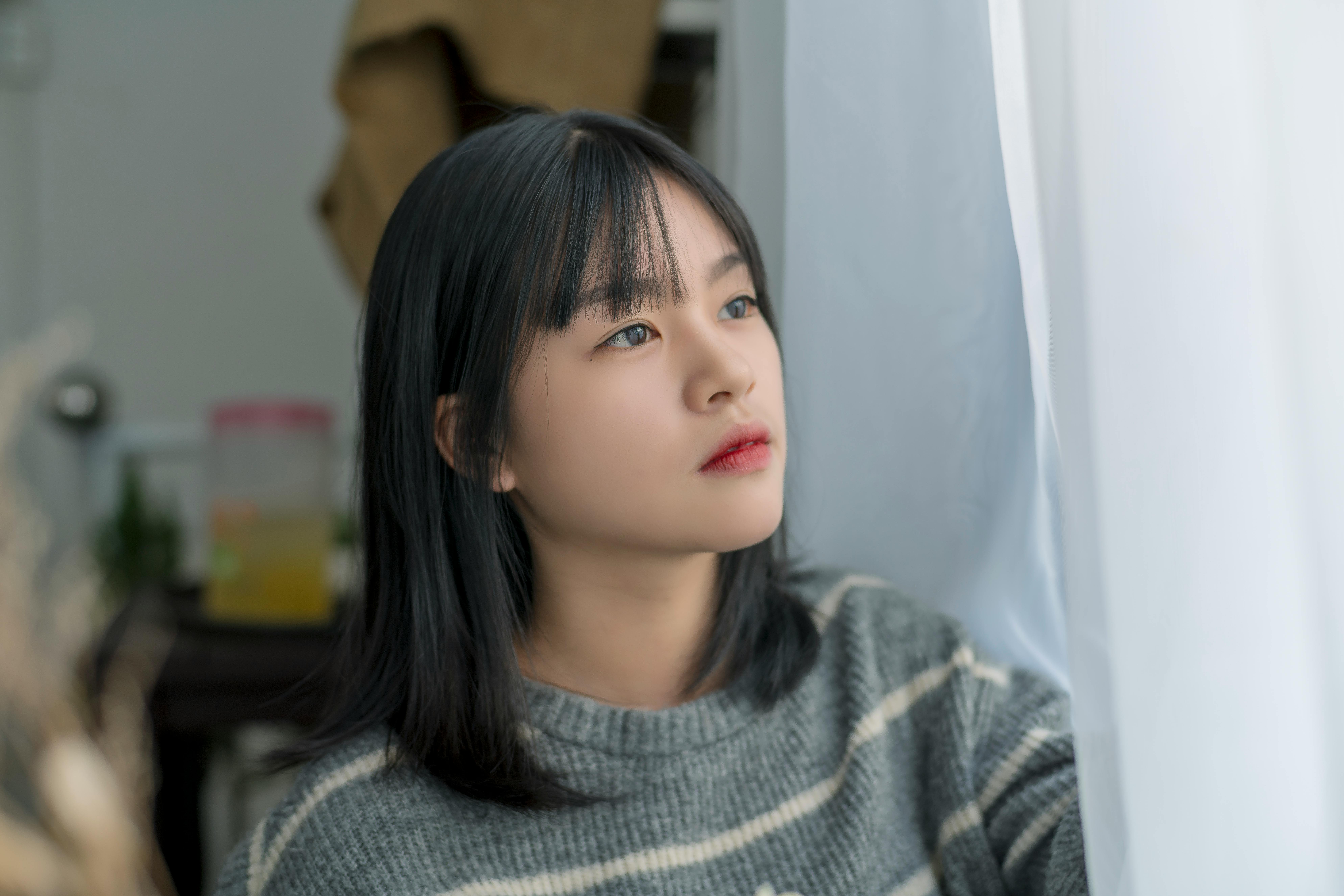 Young woman in a striped sweater gazing thoughtfully out of a window with soft indoor lighting.
