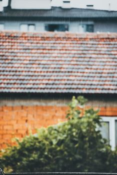 Blurred view of rain falling on a reddish-tiled roof with green bush foreground, evoking a serene rainy day.