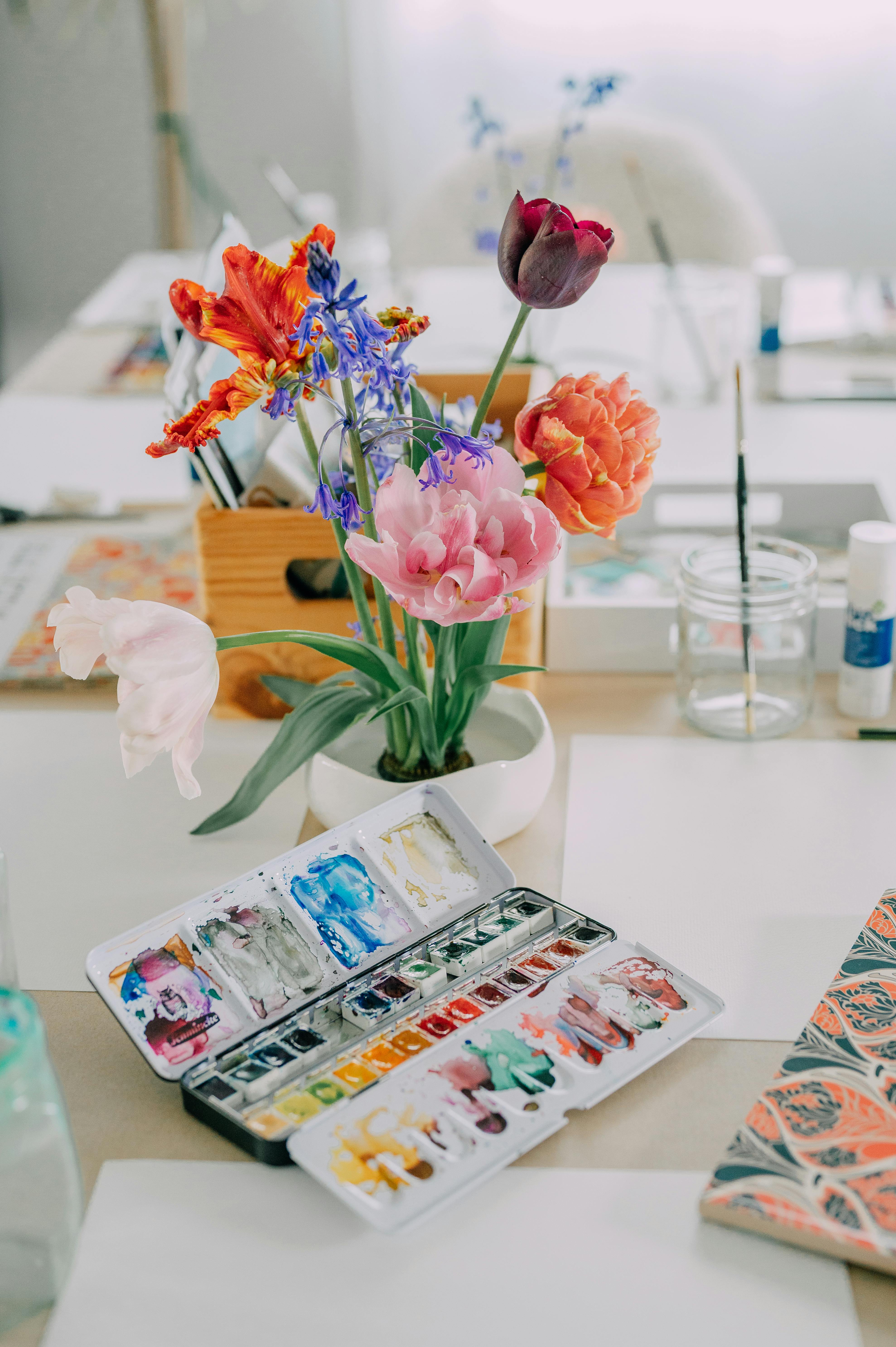 Colorful flowers and open watercolor palette on an art studio workspace.