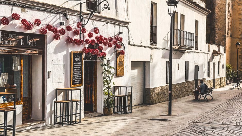 Escena callejera soleada en Cádiz, España, que muestra una pintoresca tienda con decoraciones festivas y una persona sentada en un banco.