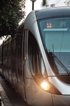 Sleek silver tram with glowing headlights traveling along city street under trees.