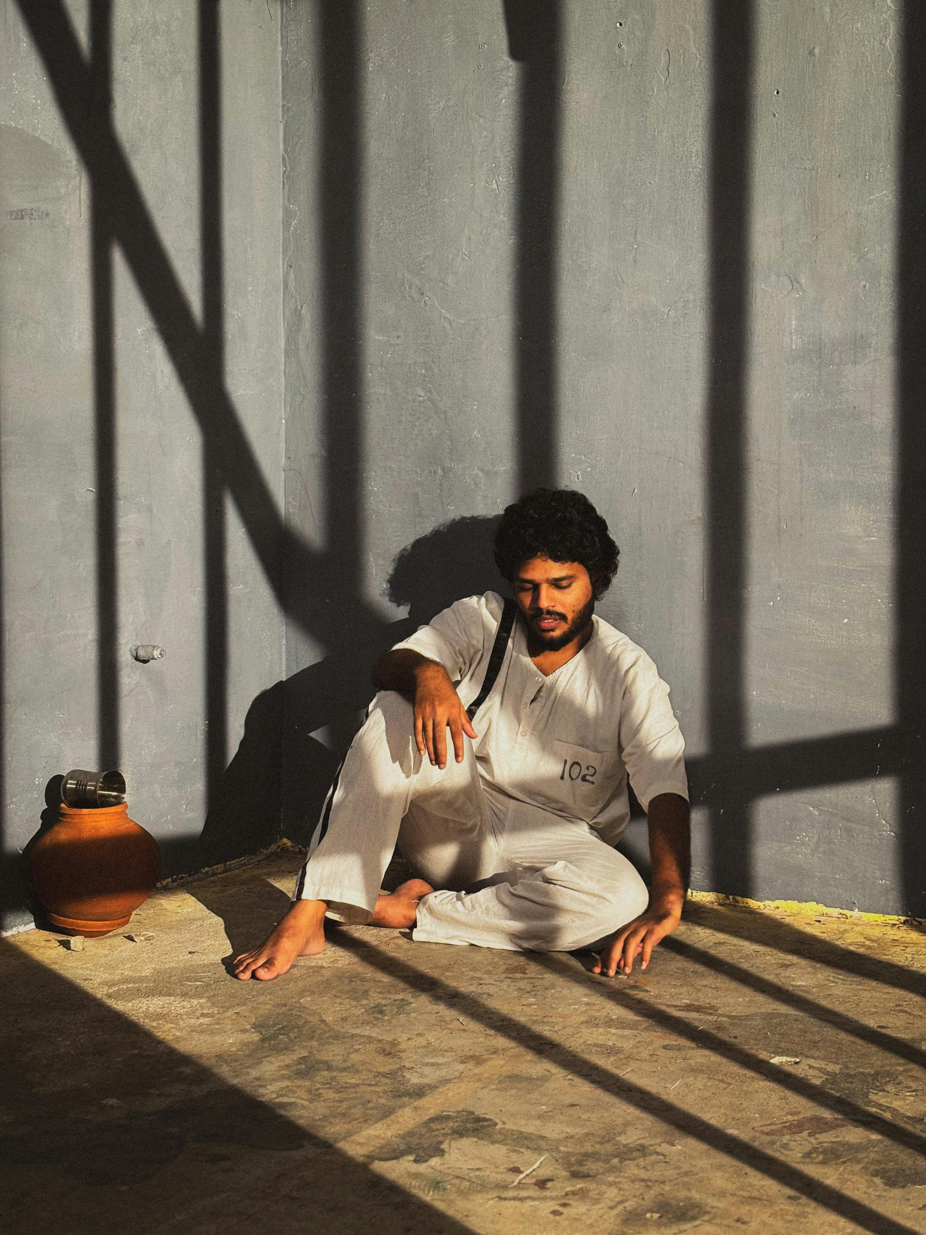 grátis Um homem de uniforme branco está sentado no chão de uma prisão iluminada pelo sol, projetando sombras. Foto profissional