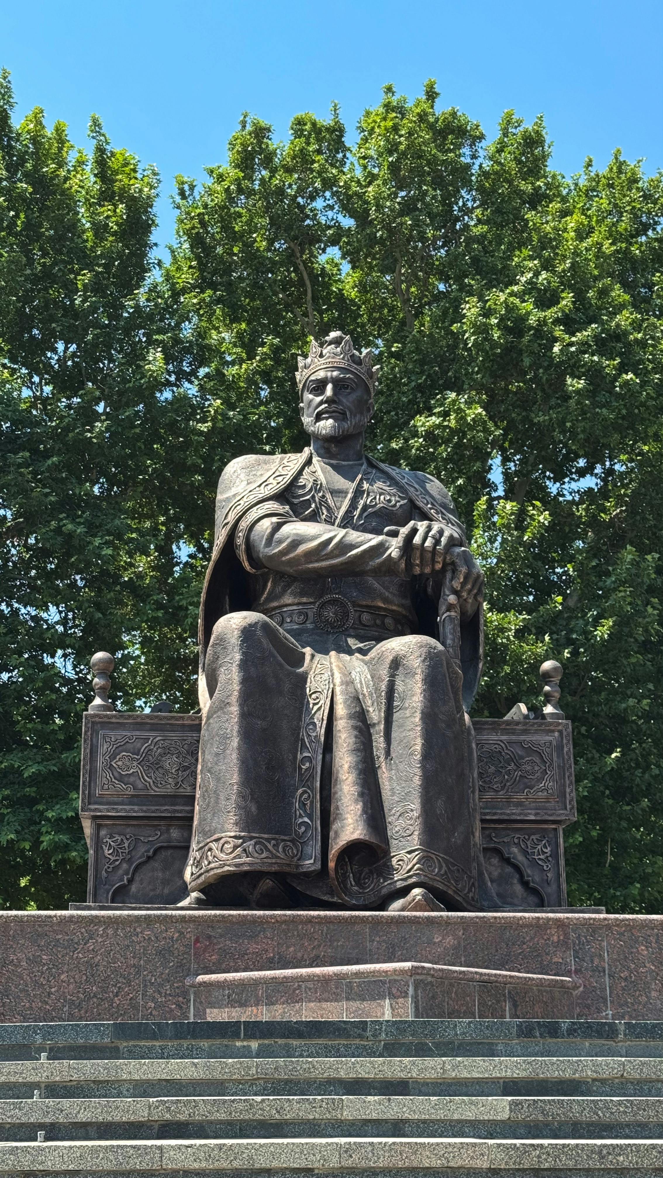 Bronze statue of seated monarch surrounded by lush trees under clear blue sky.