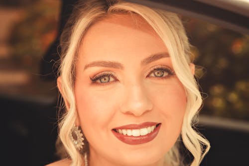 A close-up shot capturing a smiling blonde woman with vibrant makeup and earrings.