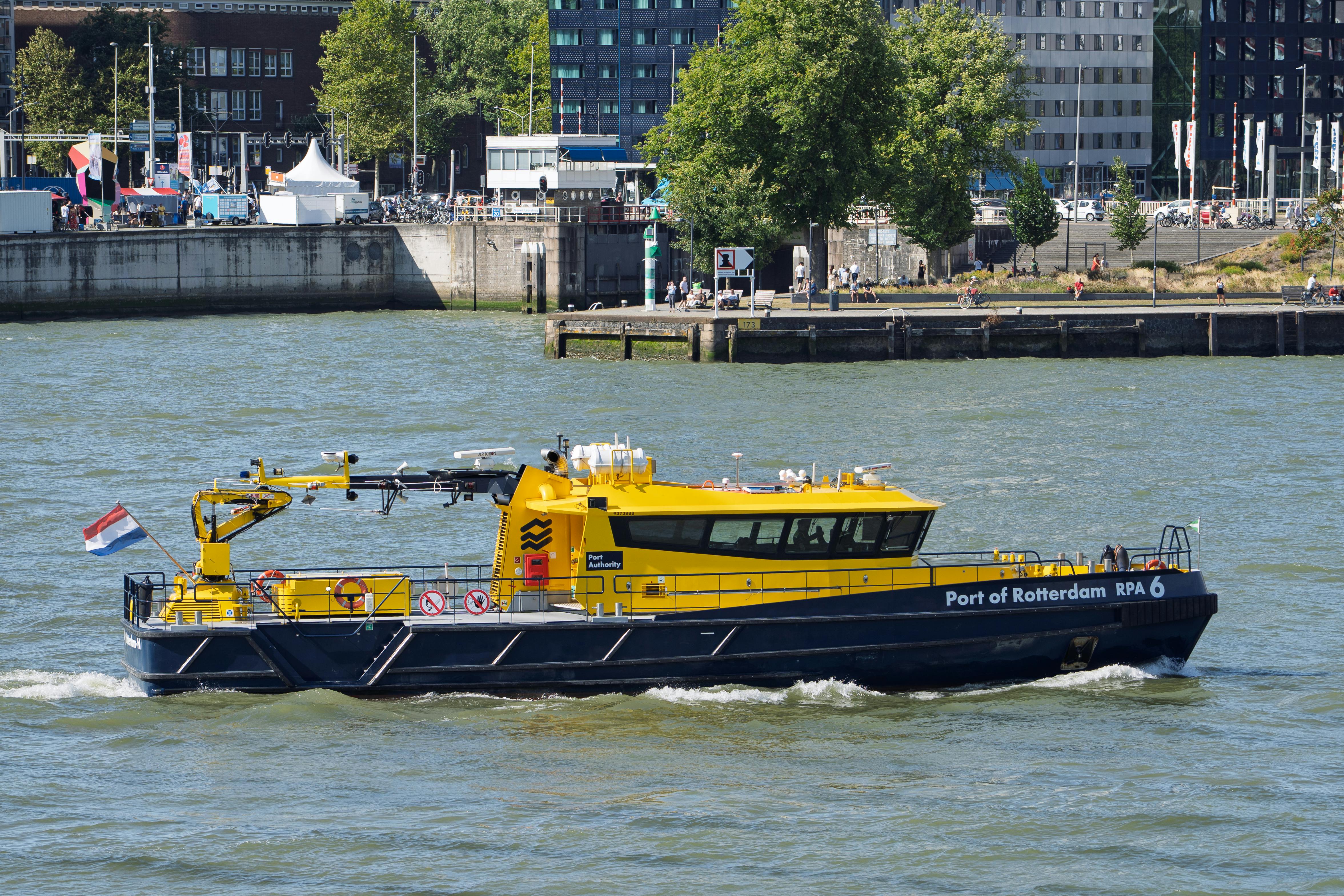 A Port of Rotterdam patrol boat cruises along a busy urban riverbank on a sunny day.