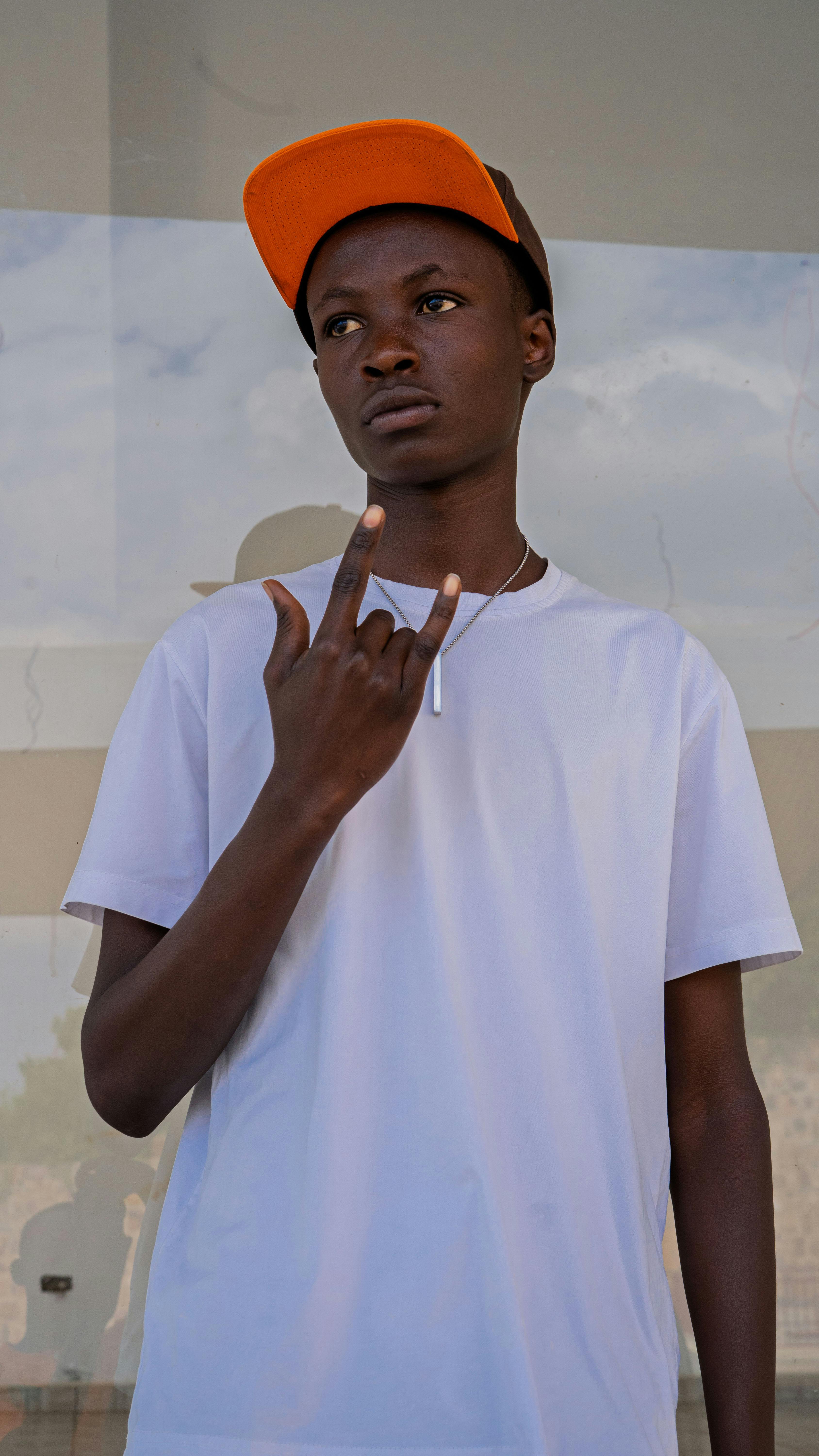 Teenager in white t-shirt and orange cap making a gesture outdoors.