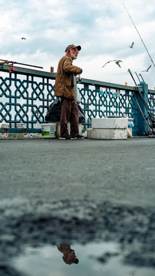 Free Elderly man engages in fishing on an urban bridge. Seagulls in sky create a picturesque scene. Stock Photo