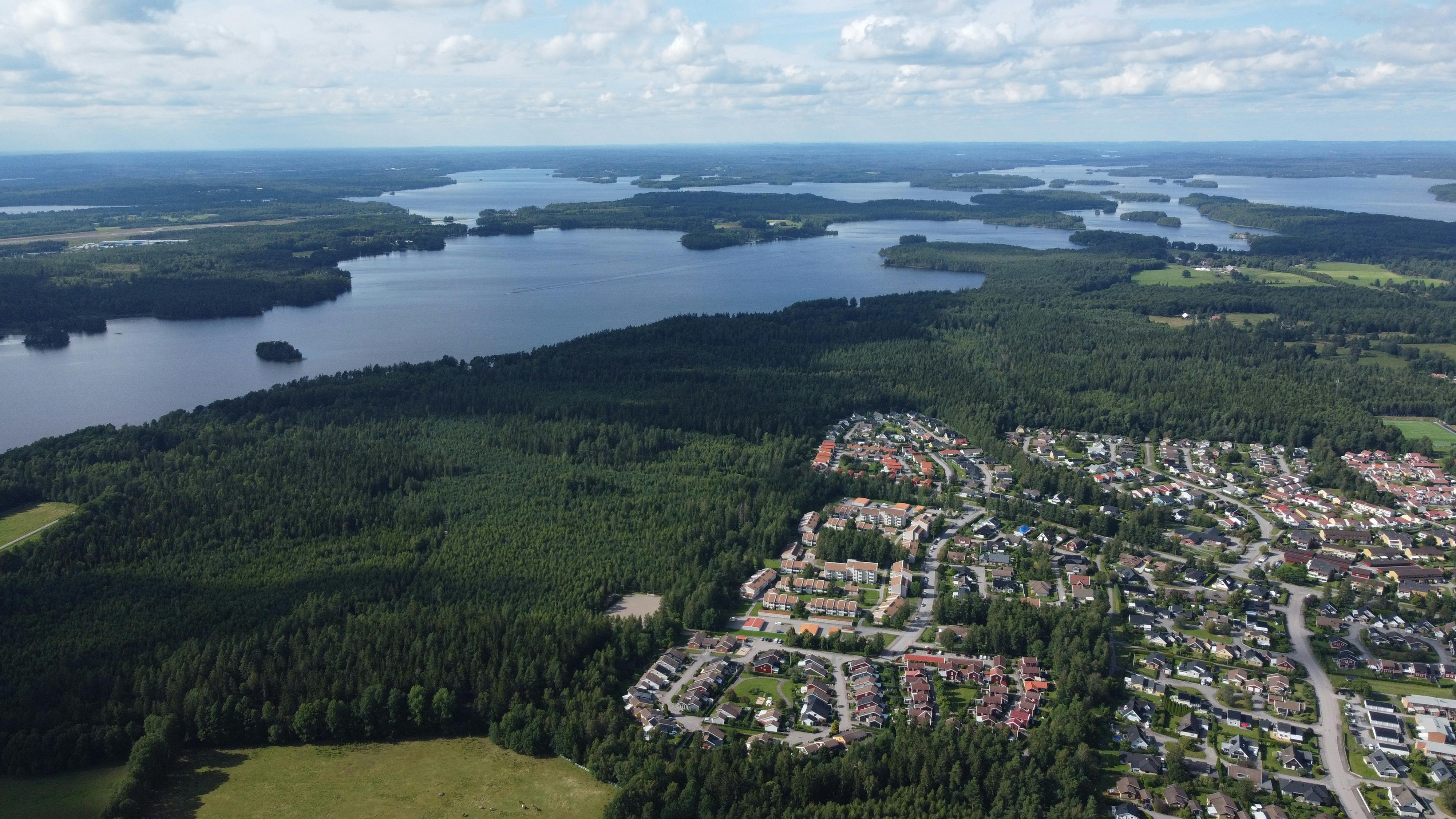Aerial view of Swedish landscape with lush forests, serene lakes, and suburban areas under a blue sky.