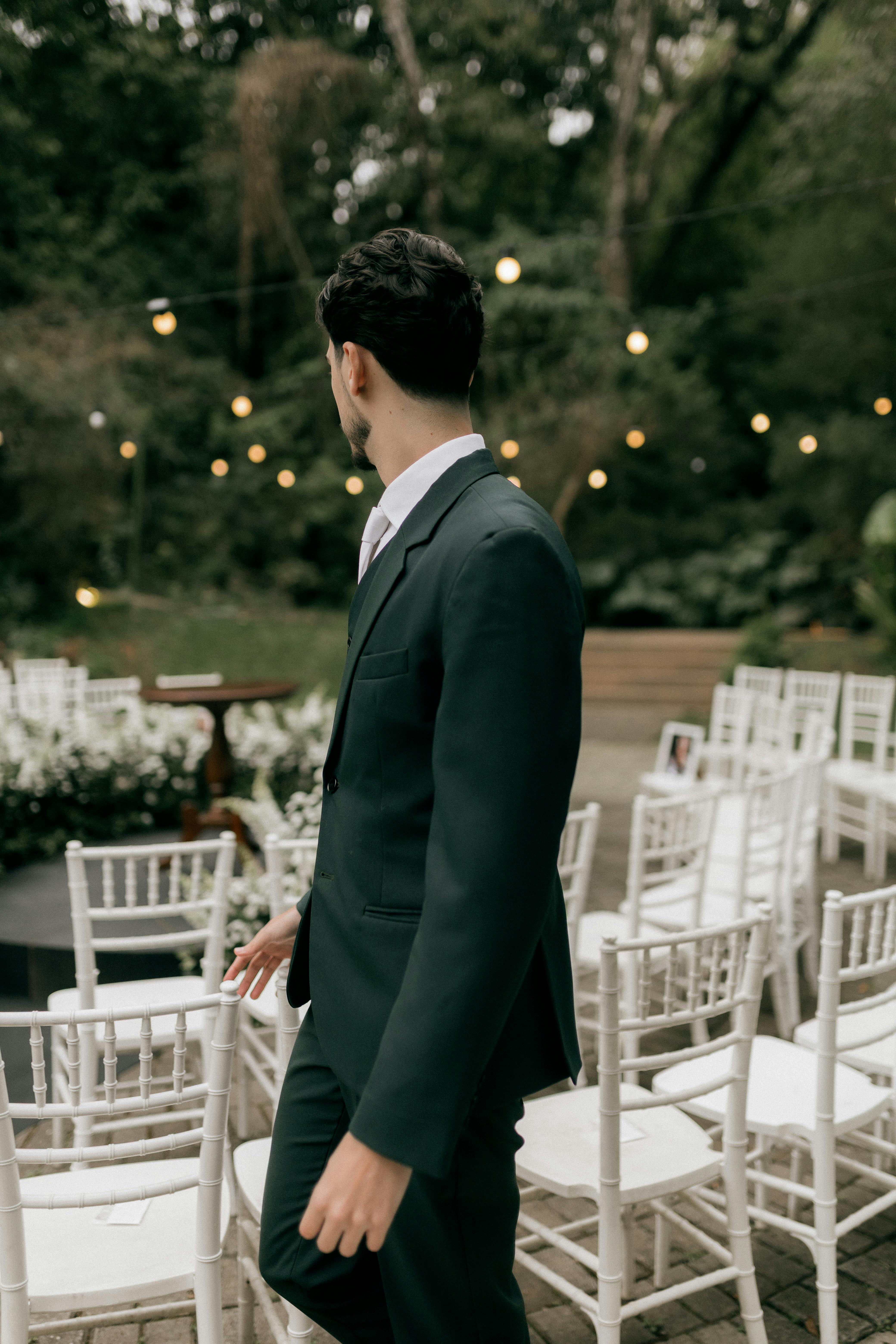 Man in a formal suit at an outdoor wedding venue with chairs and string lights.