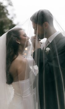 A tender moment between a bride and groom under a wedding veil outdoors.