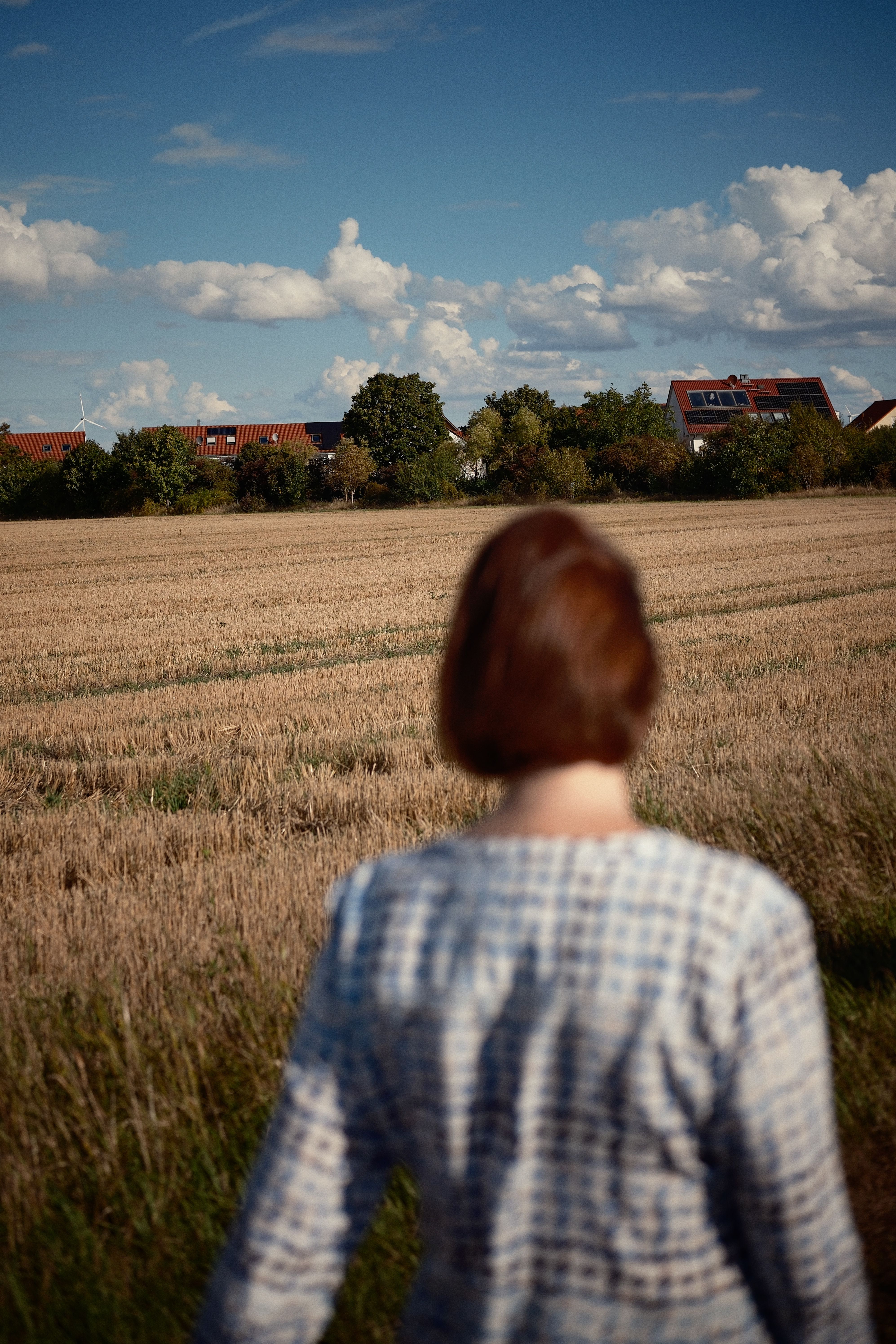 A woman stands in a field gazing towards a scenic rural landscape under a cloudy sky.