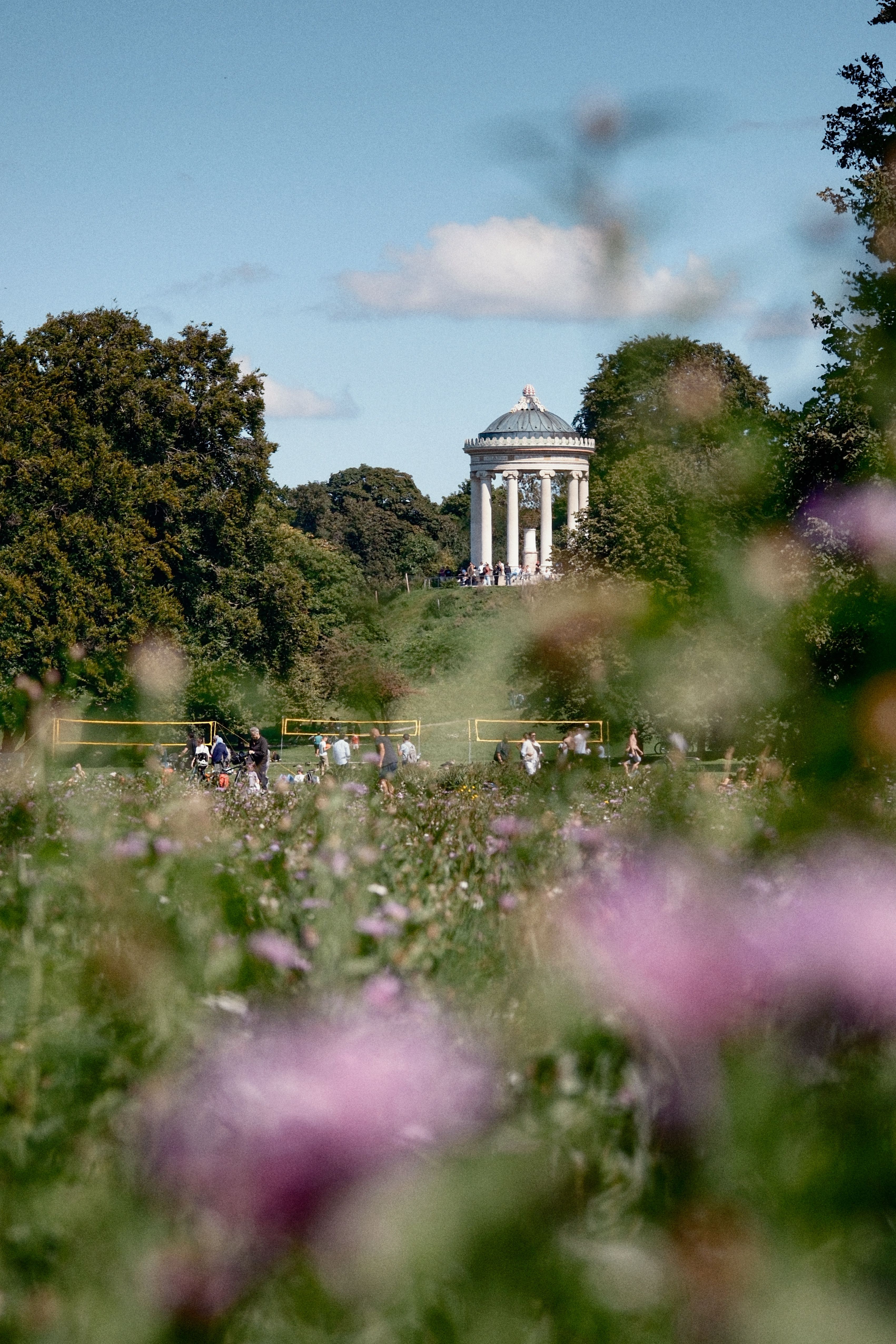 A distant view of the Temple of Apollo surrounded by lush greenery and blooming flowers on a sunny day.