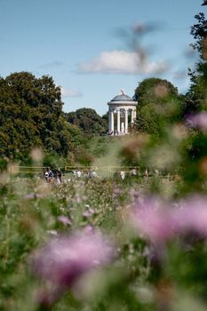 A distant view of the Temple of Apollo surrounded by lush greenery and blooming flowers on a sunny day.