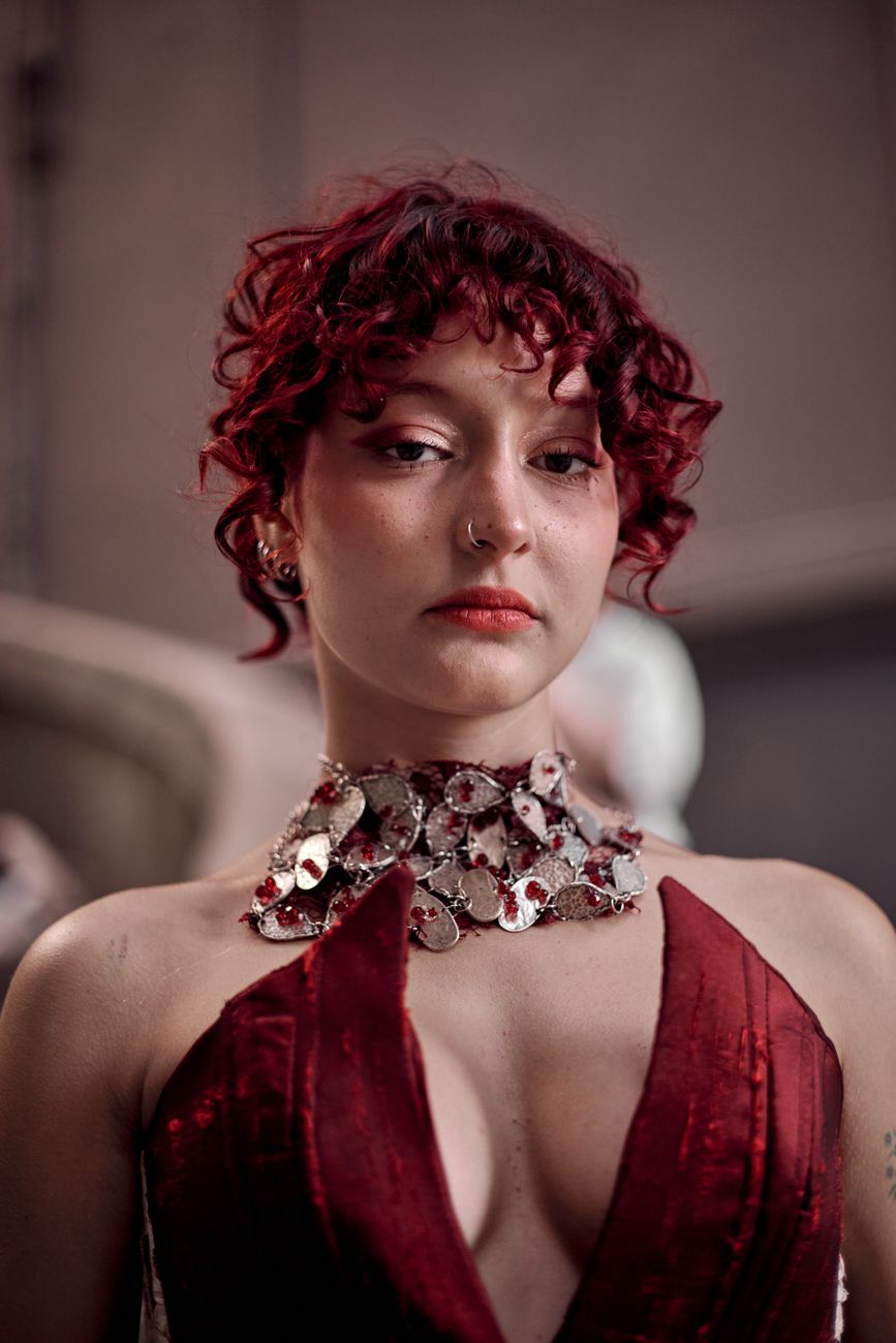 Elegant portrait of a woman with red curly hair in a luxurious red dress and statement necklace.