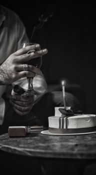 Black and white image of a hand holding a drink beside a cake with a candle, evoking a classic celebration mood.