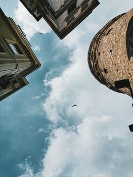 Low angle view of an old tower against a cloudy sky, framed by surrounding buildings.