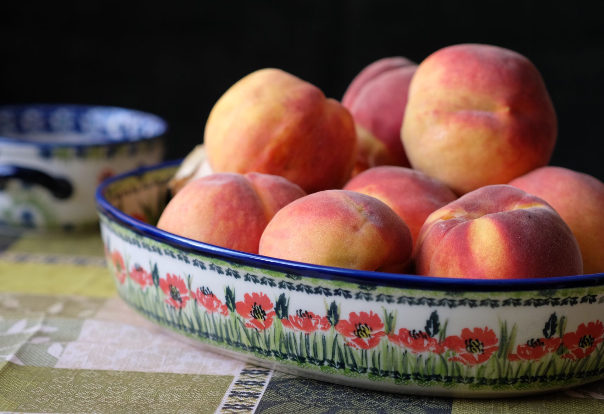 A vibrant display of ripe peaches in a floral ceramic bowl set on a patterned tablecloth.