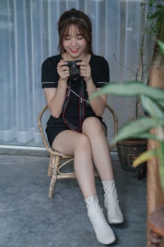 Smiling woman seated indoors, holding a camera with a thoughtful expression.