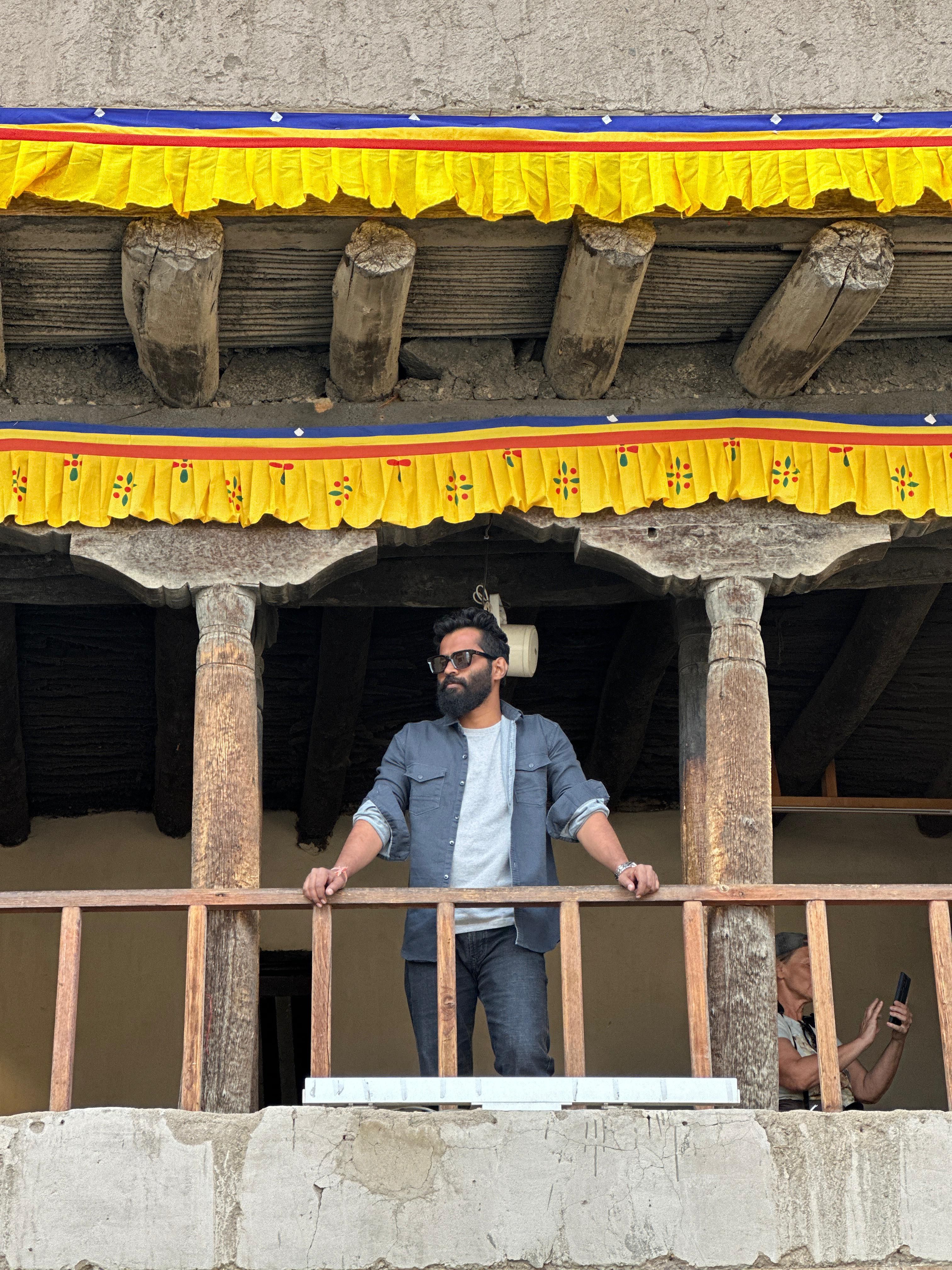 Man standing on a monastery balcony in Leh, showcasing traditional architecture and vibrant textiles.