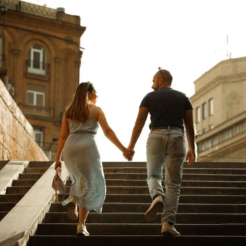Free A romantic couple holding hands and walking up stone steps in Armenia, surrounded by historic architecture. Stock Photo