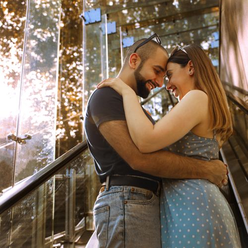 Free A young couple shares a loving embrace on a glass staircase surrounded by natural sunlight. Stock Photo