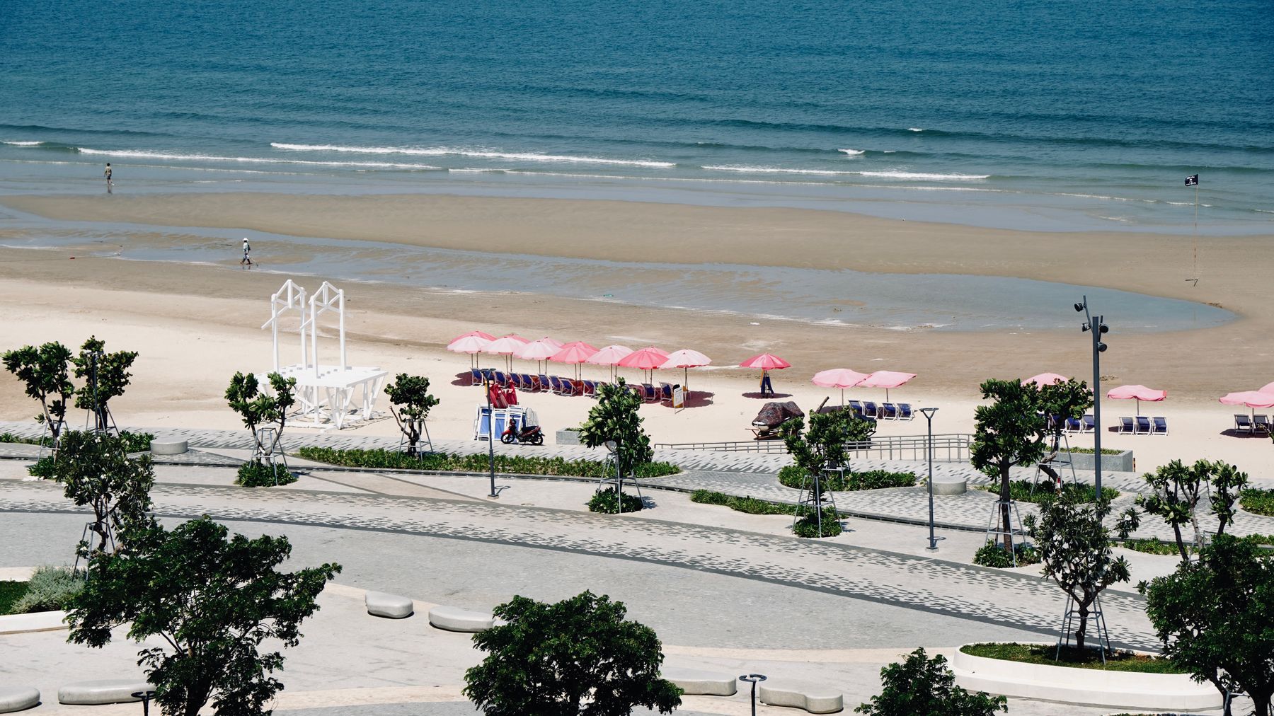 A sunny beach with pink umbrellas, clear blue ocean, and a promenade in the foreground.
