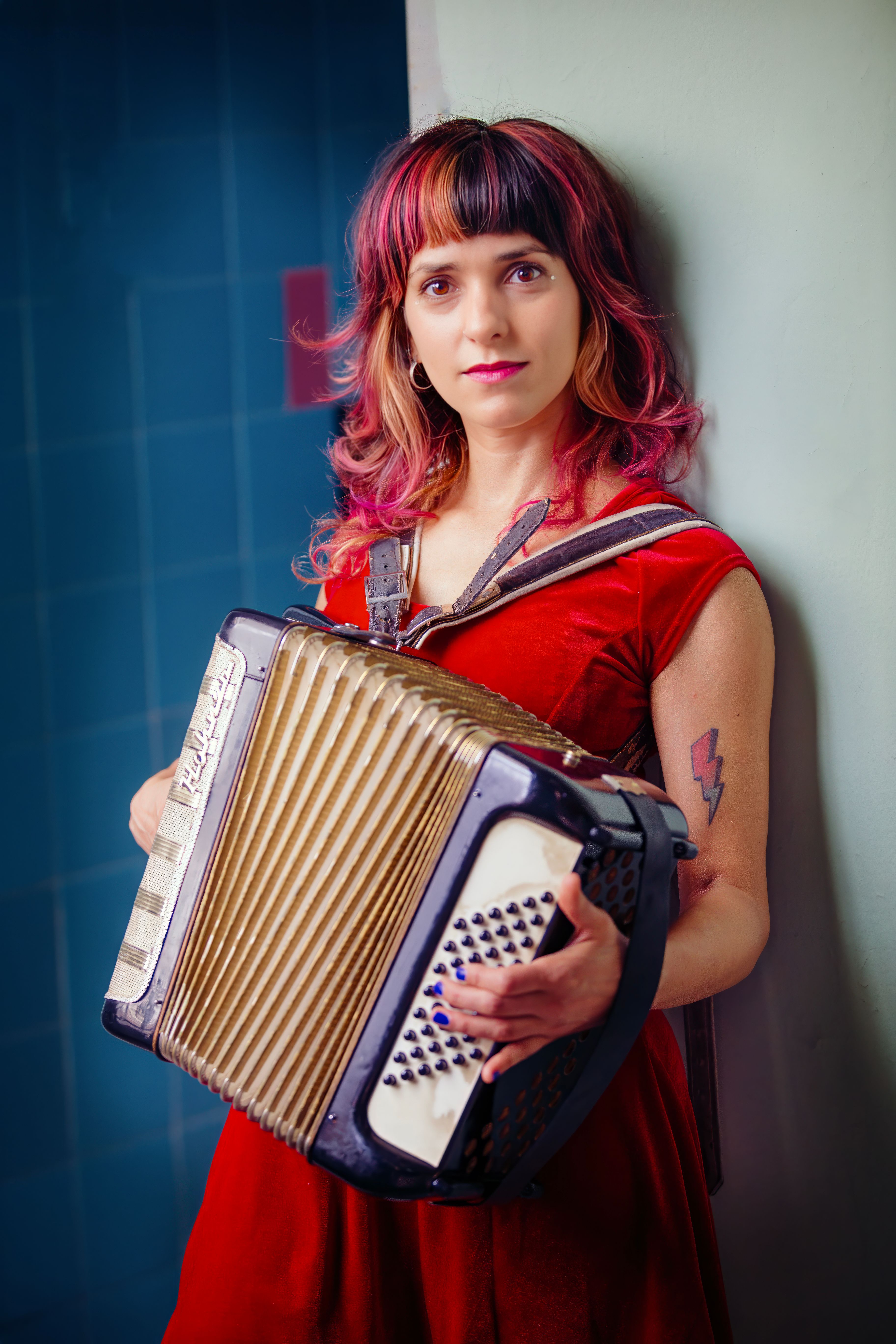 Colorful portrait of a woman with pink hair in a red dress playing the accordion indoors.