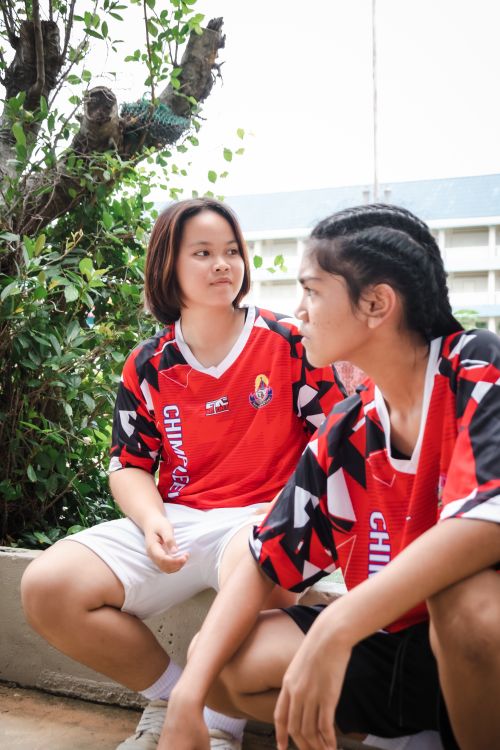 Free Two teenage girls in red soccer jerseys sitting outdoors, engaged in a conversation. Stock Photo