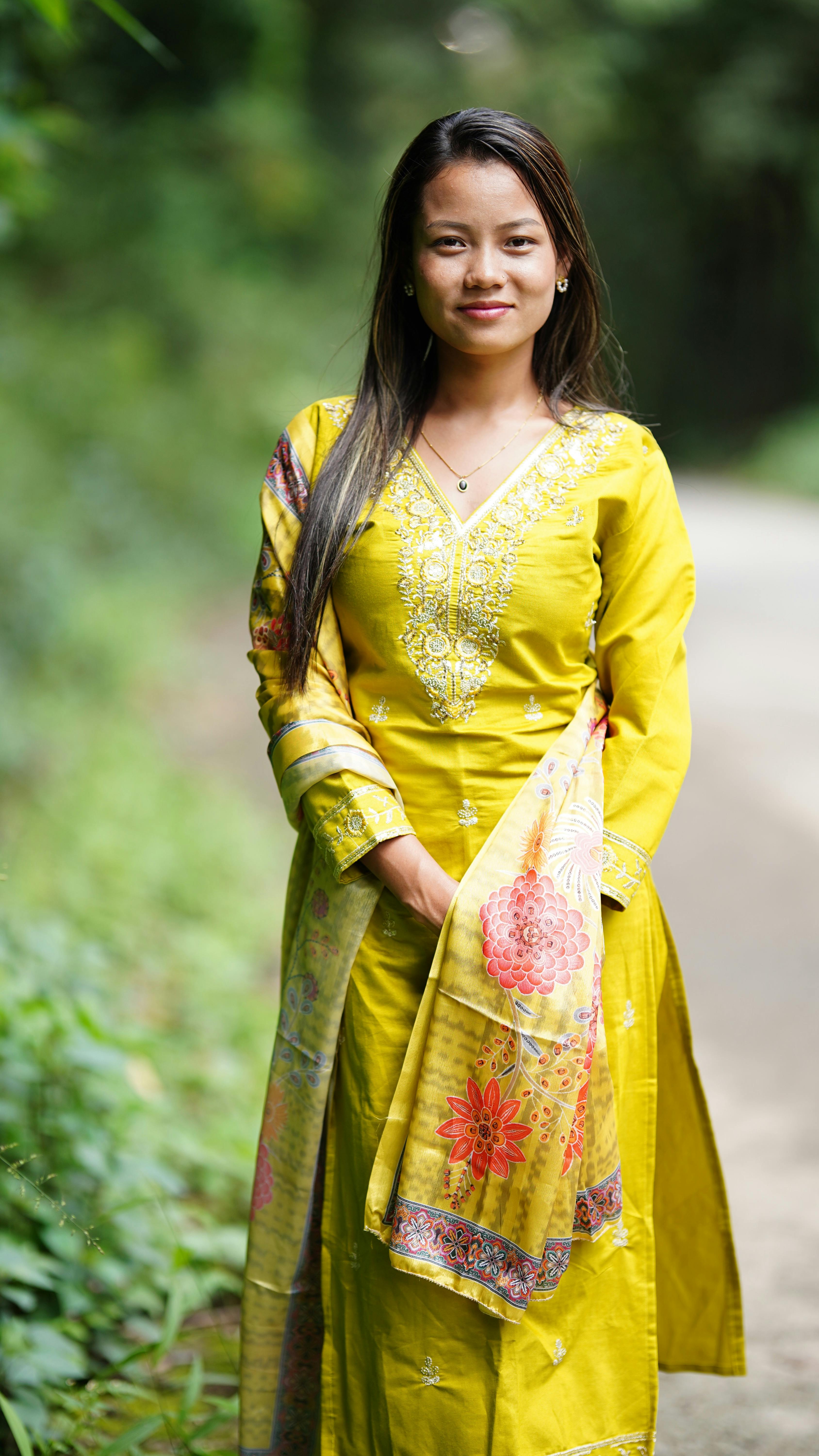A woman in a vibrant yellow traditional dress stands on a path surrounded by greenery.