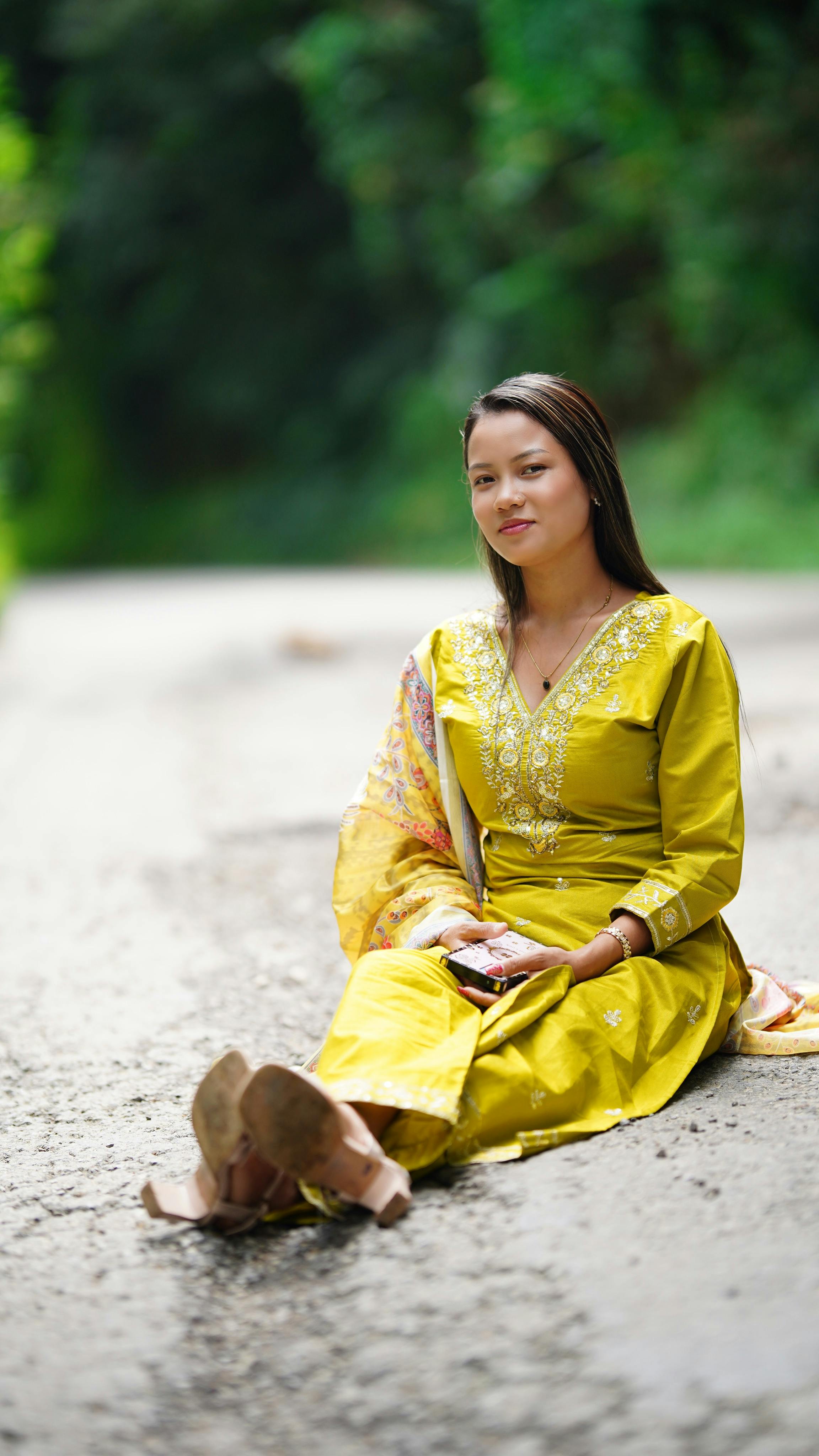 A woman in a yellow dress sits on a road surrounded by greenery, enjoying a peaceful day outdoors.