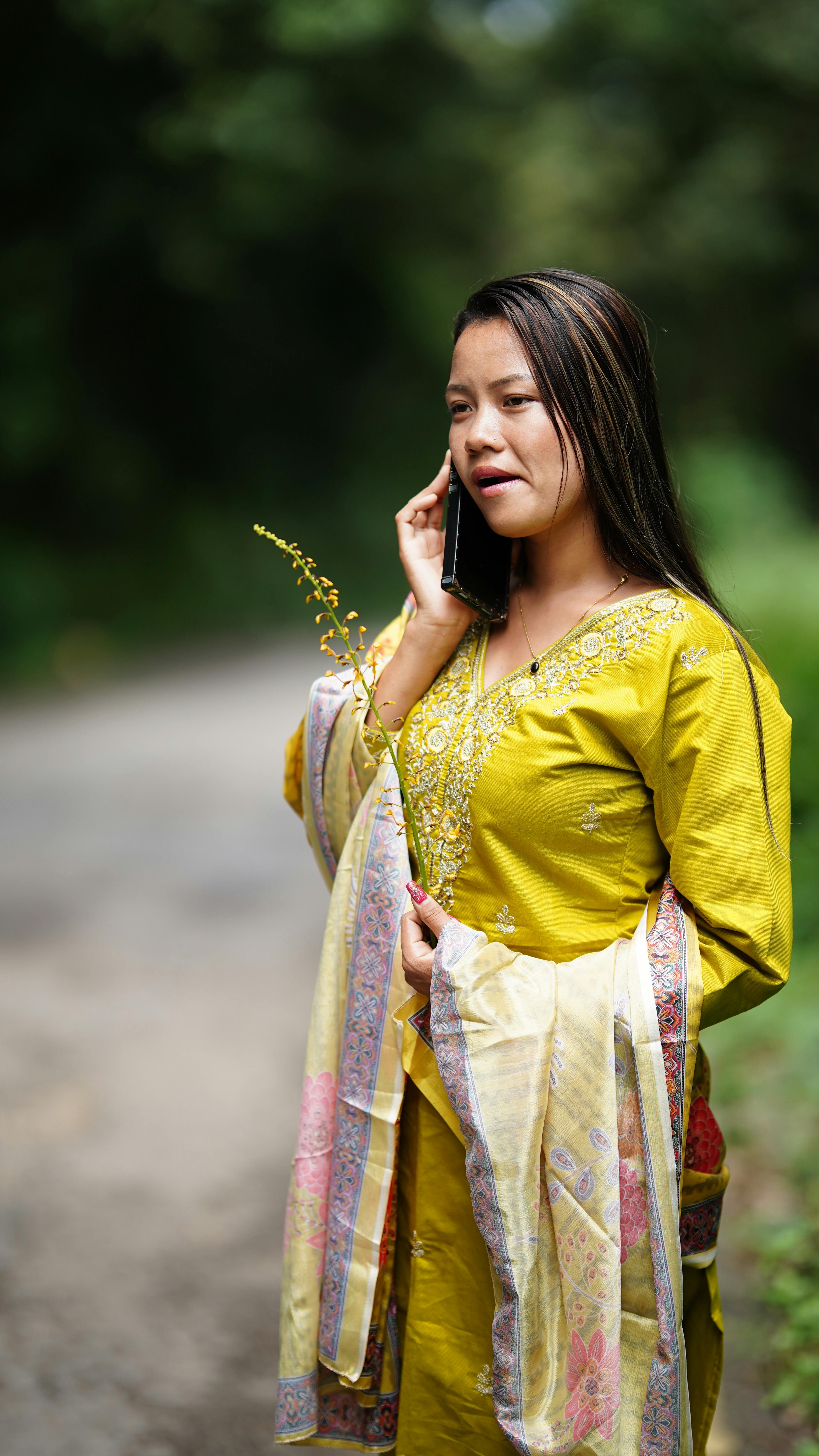 A woman in yellow traditional attire speaks on the phone while walking outdoors.