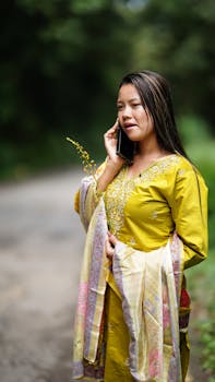 A woman in yellow traditional attire speaks on the phone while walking outdoors.
