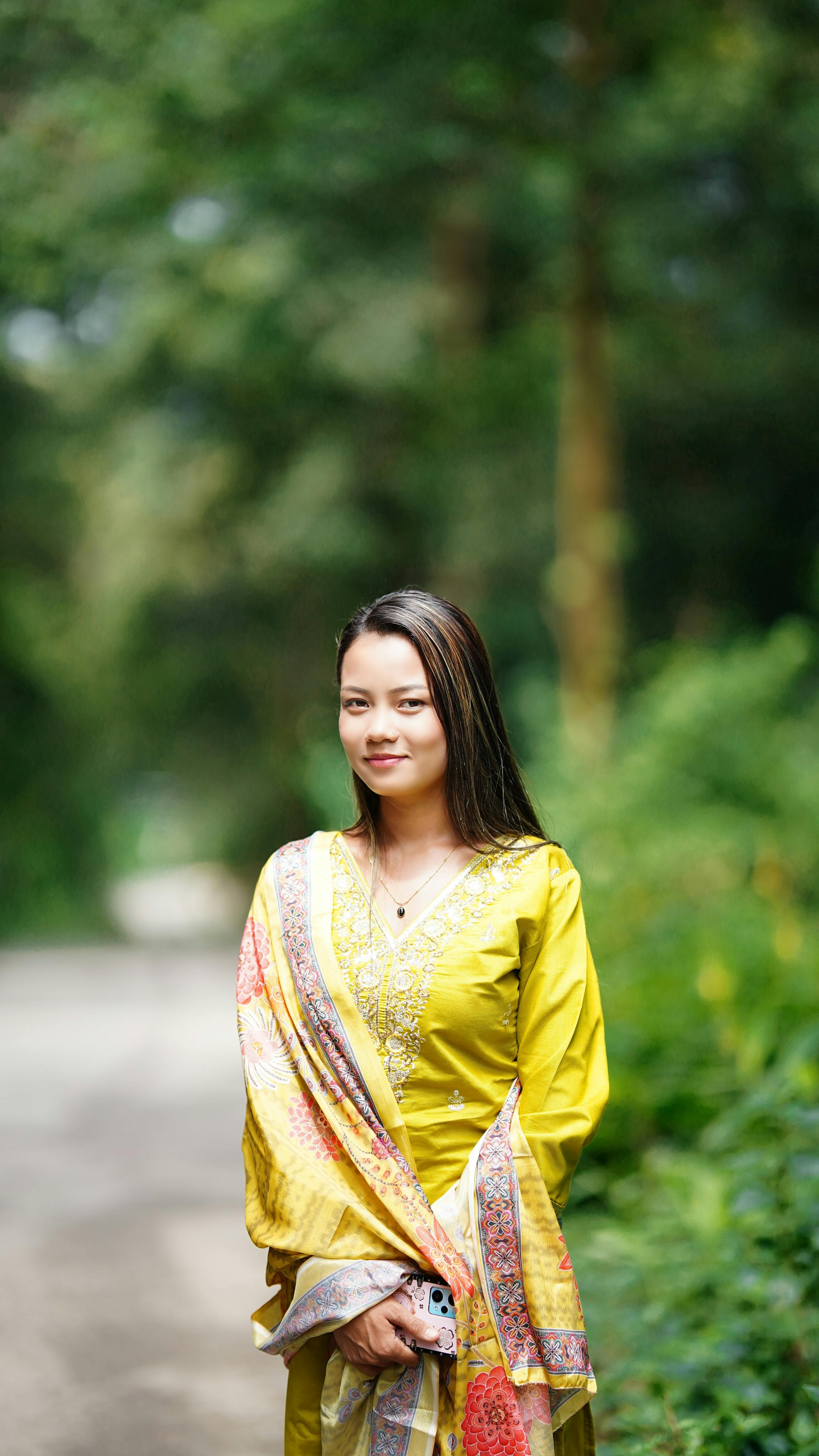 Smiling woman in a yellow traditional outfit standing in a lush green outdoor setting.
