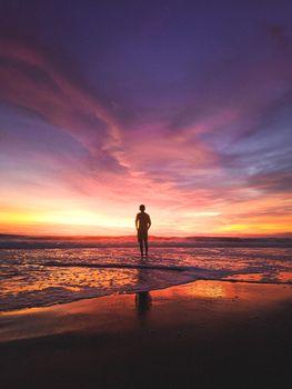 A lone figure stands on a beach, silhouetted against a dramatic sunset with vivid colors.