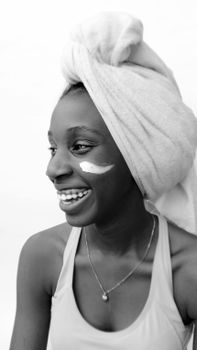 Black and white portrait of a smiling woman with skincare cream and towel on head.