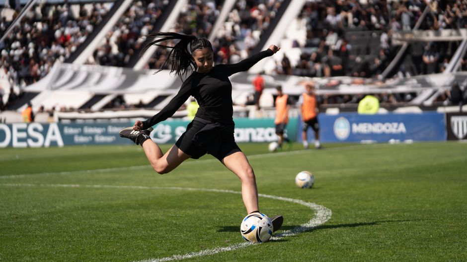 A female soccer player powerfully kicks a ball during warm-up on a sunny day.