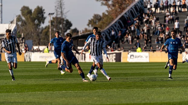 Football players in action during a sunny outdoor match with a vibrant crowd.