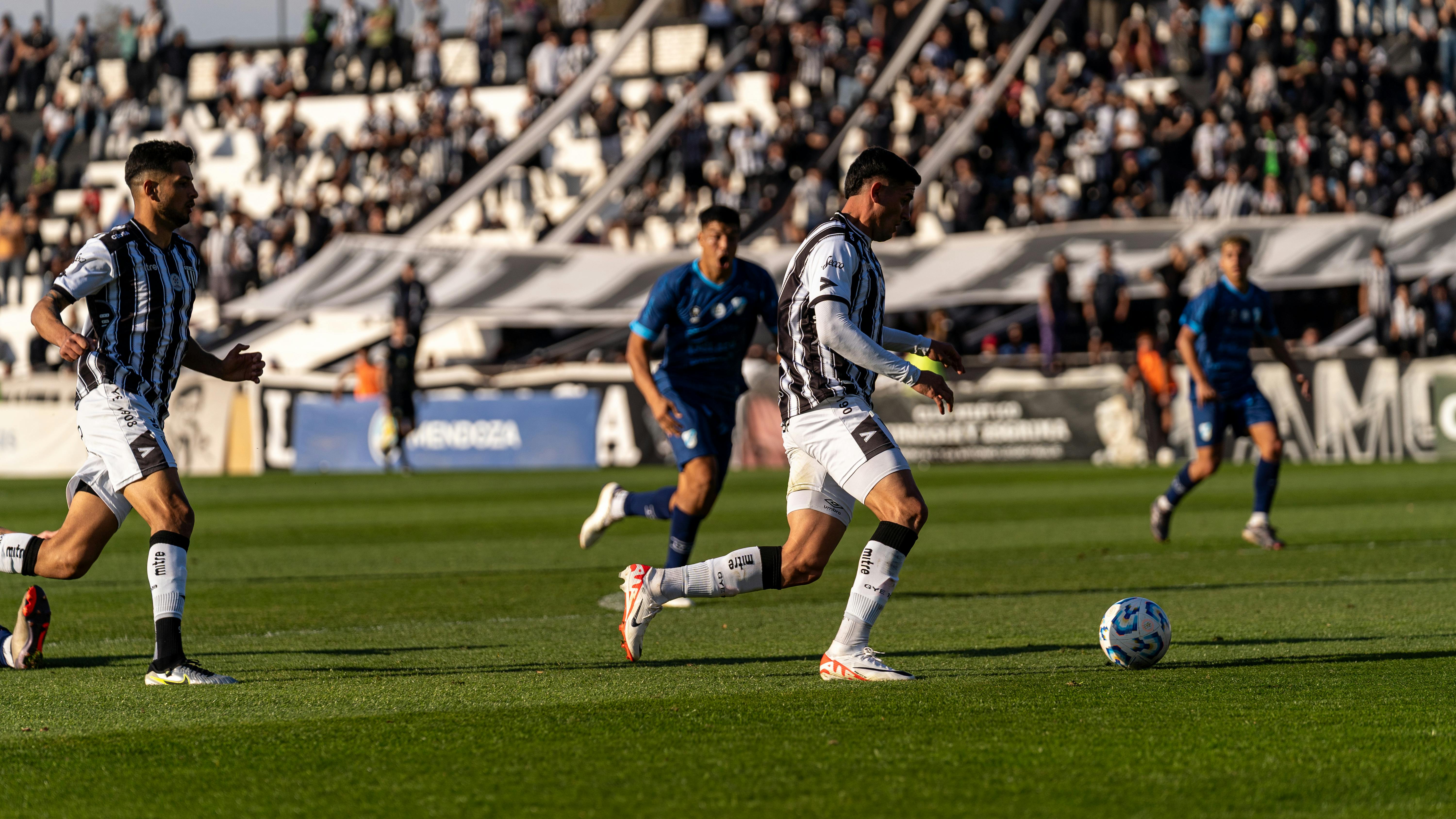 Soccer players in action during a lively match on a sunny day with a cheering crowd.