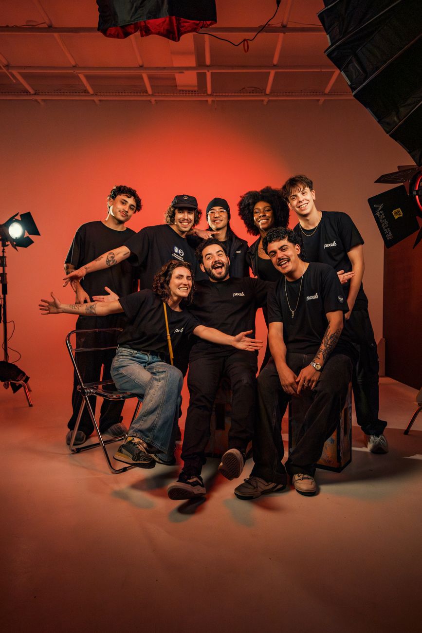 Un groupe de huit jeunes adultes souriants pose ensemble dans un studio professionnel avec un fond rouge et un éclairage de plateau. Certains sont assis, d'autres debout, portant des t-shirts noirs.