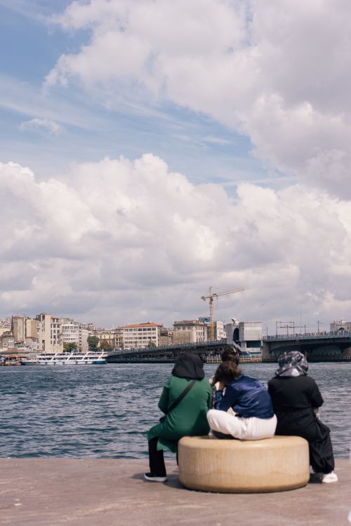 Free Three women sit by the water with a view of Istanbul's skyline under a bright sky. Stock Photo