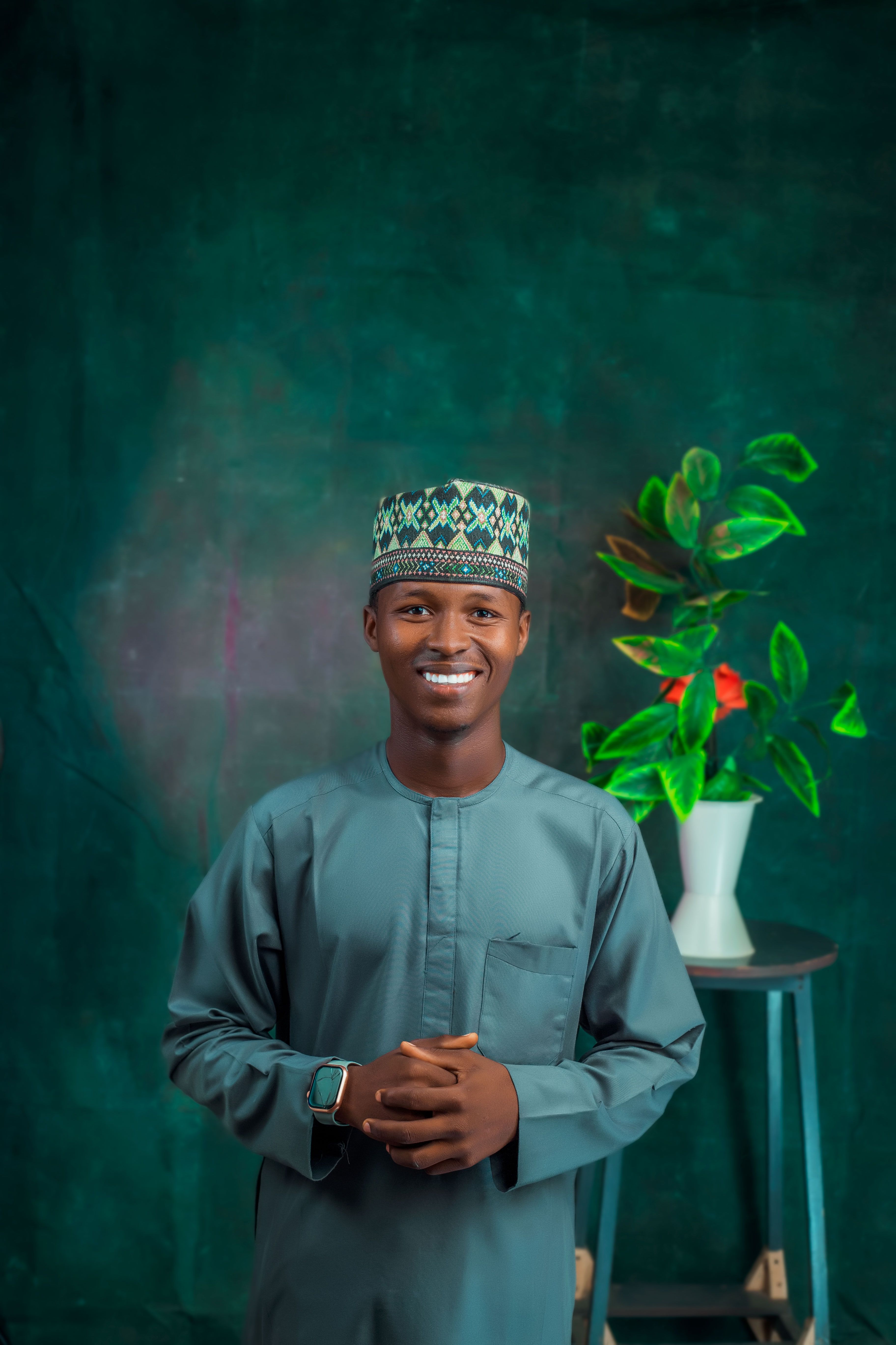 A cheerful man in traditional attire stands indoors with a vibrant plant nearby.