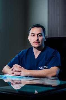 Confident male doctor in blue scrubs seated at a reflective desk in an office setting.