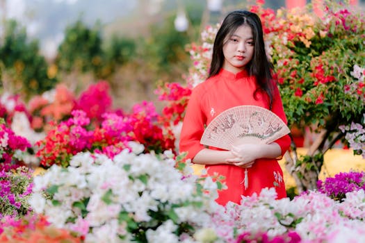 Young woman in a vibrant red dress holding a fan amidst a colorful flower garden.