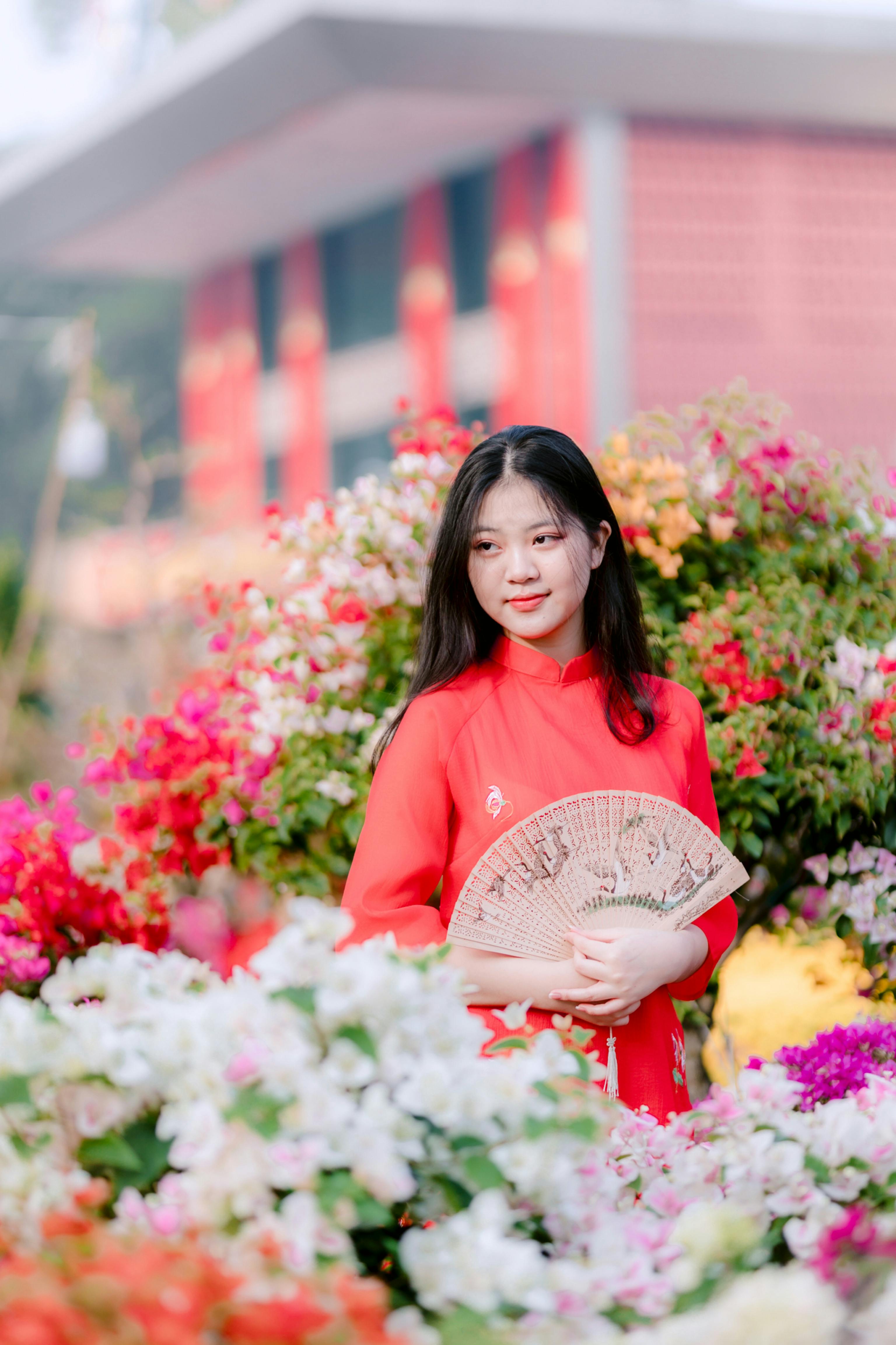 Portrait of a young woman in red holding a fan surrounded by vibrant flowers outdoors.