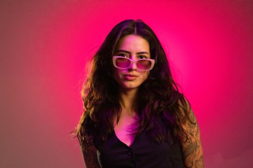 Free Portrait of a stylish woman with tattoos wearing pink sunglasses, against a vibrant pink background. Stock Photo