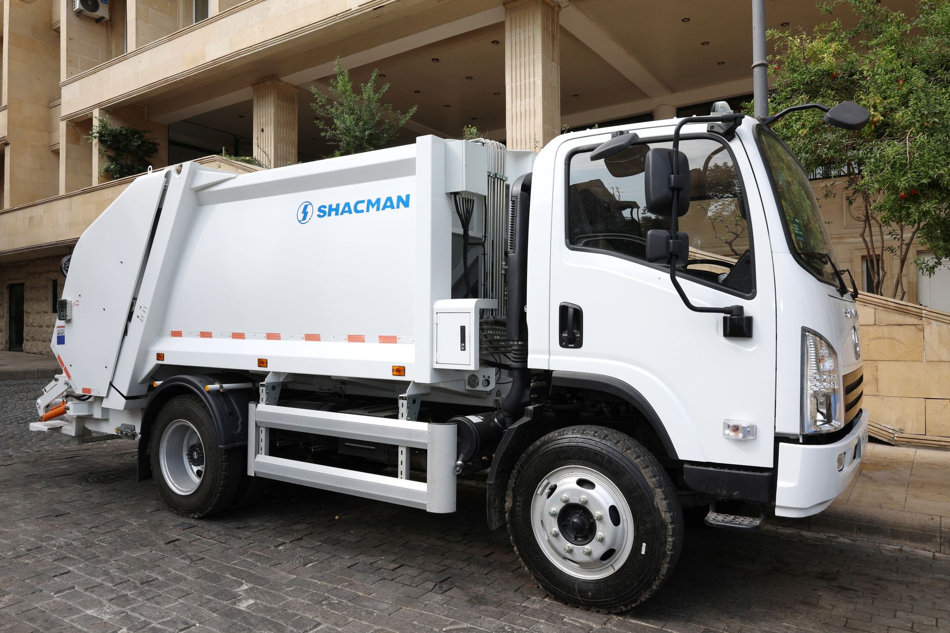 White Shacman garbage truck parked on a cobblestone street in an urban area, ready for waste collection.