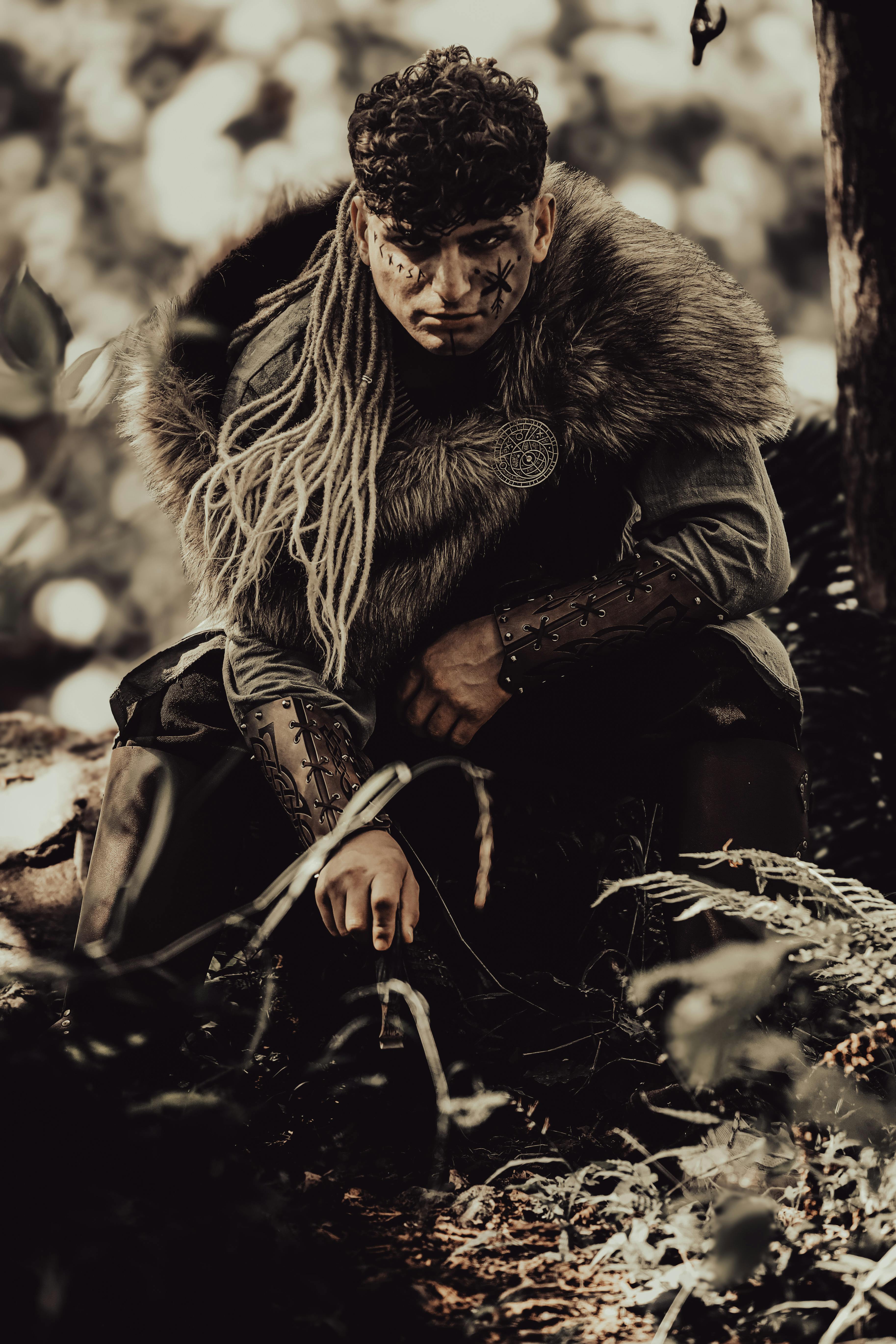 A warrior with tribal markings poses in a dense, mysterious forest, evoking an ancient time.