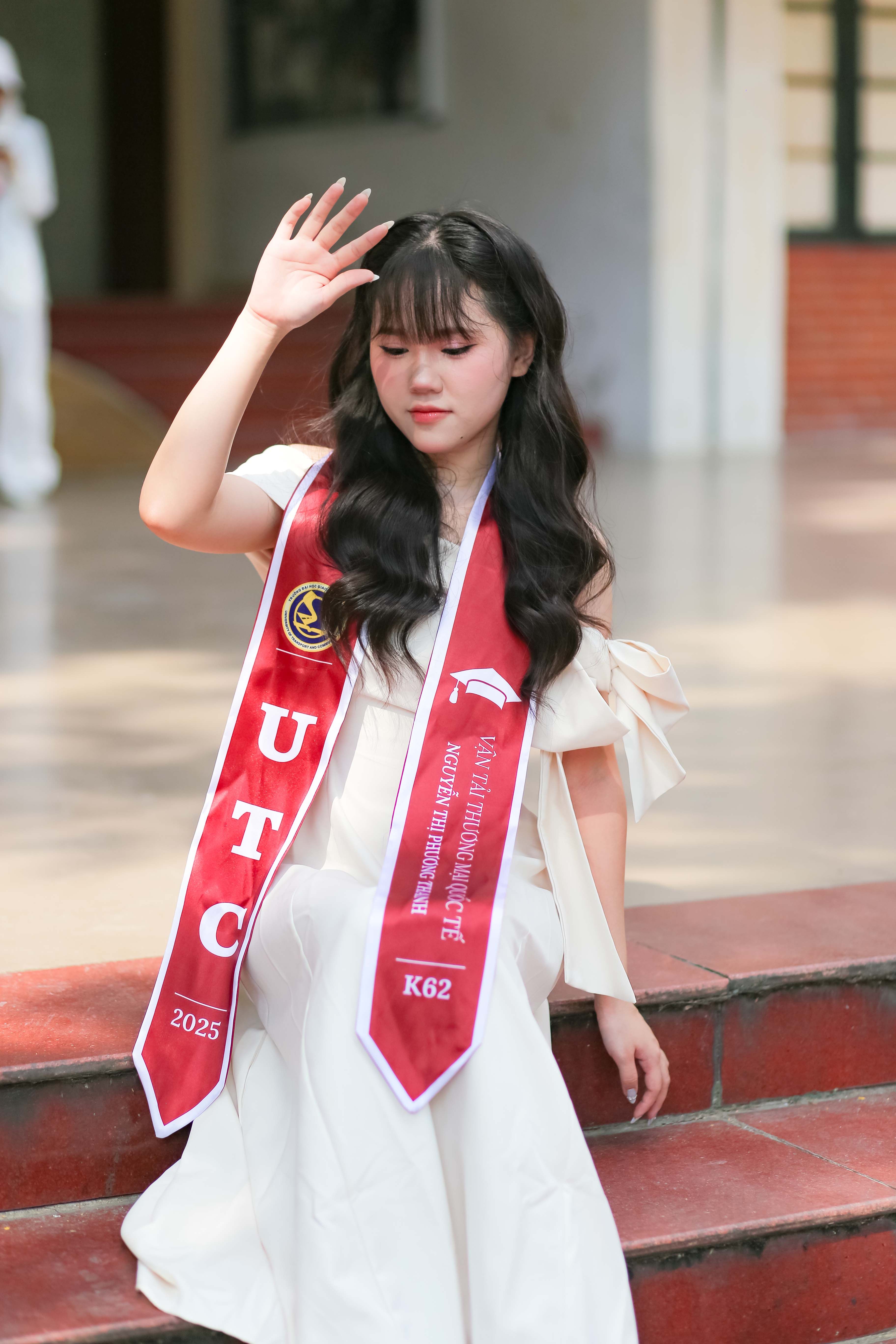 Portrait of a young woman in graduation attire raising her hand outdoors.