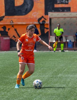 Female athlete in orange uniform dribbling a soccer ball during a daytime match on an outdoor field.