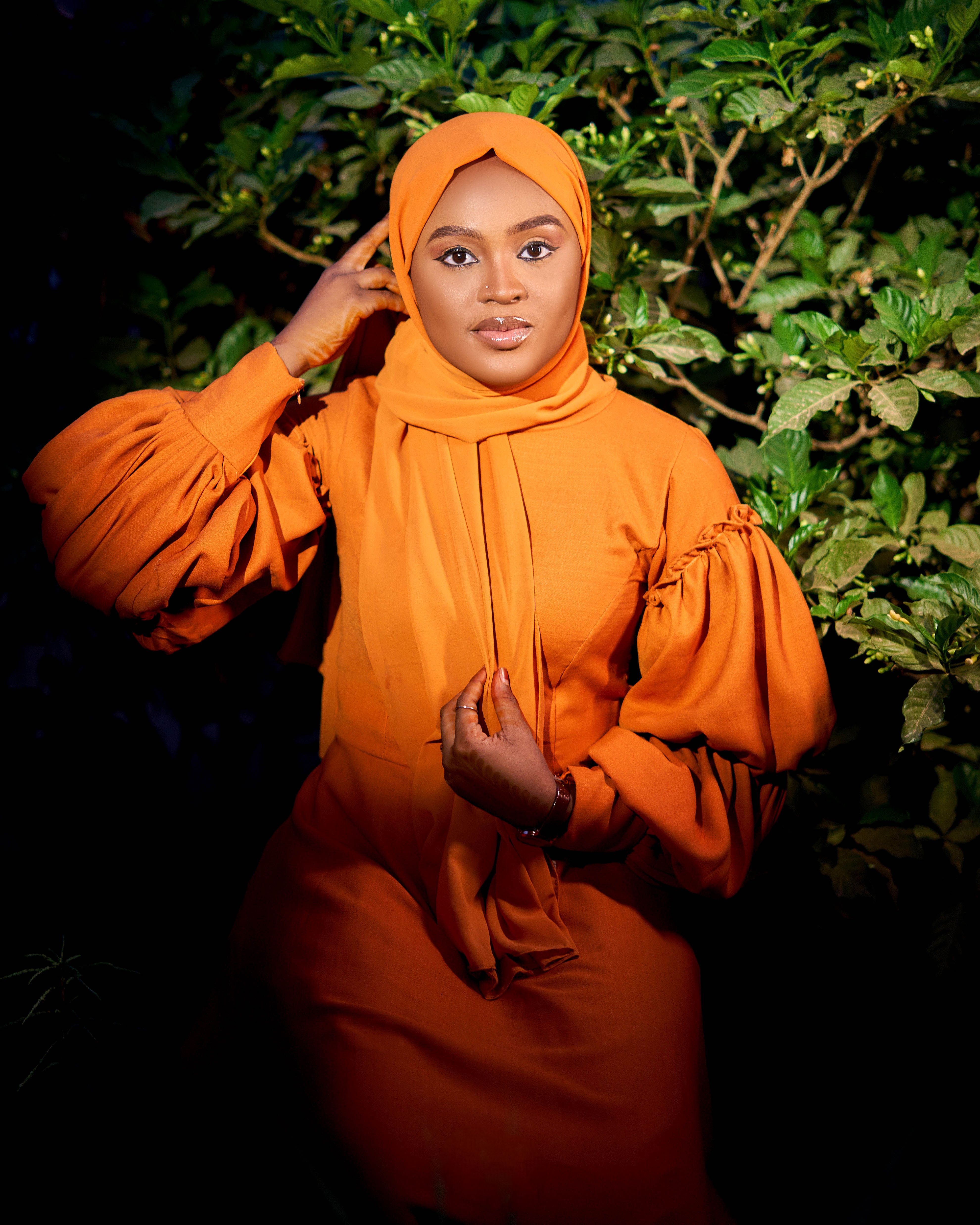 A beautiful woman in an orange abaya poses confidently against a lush green backdrop, exuding elegance and grace.